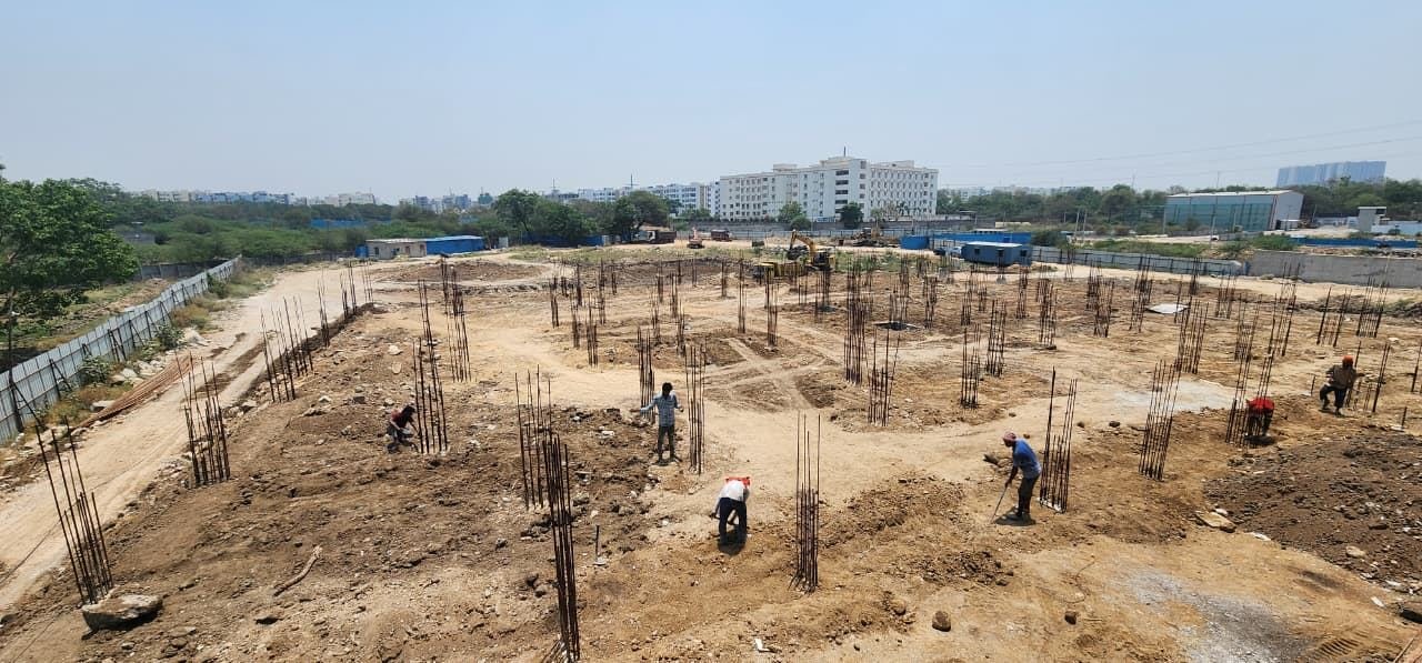 On-site photograph of the Fortune Towers foundation stage: rows of vertical steel rebar columns rising from cleared, levelled earth across a wide plot, with workers laying out column positions and an excavator and site sheds visible in the background, apartment buildings on the skyline beyond the boundary wall.