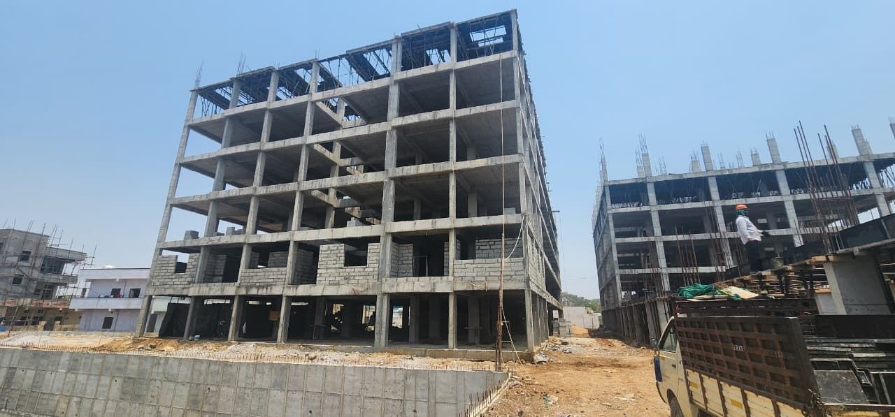 On-site photograph of a Fortune Towers tower under construction: a multi-storey R.C.C. framed structure with exposed concrete columns and brick infill walls rising six-plus levels, a second building under similar construction to the right, a loaded truck and scaffolding in the foreground.