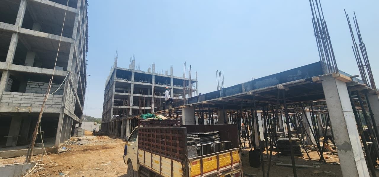 On-site photograph at Fortune Towers: a multi-storey R.C.C. framed building under construction on the left, a second structure with shuttering and props for a slab pour on the right, a material-laden truck and scaffolding props in the foreground.